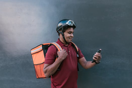 A delivery courier wearing a helmet and backpack checks his smartphone against a dark background.