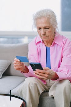 Senior woman using a credit card and smartphone for online shopping in a bright living room.